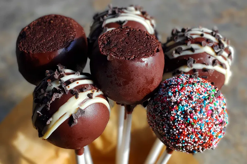 Ingredients for chocolate cake pops including chocolate cake mix, frosting, candy melts, cake pop sticks, and colorful decorating supplies arranged on a baking counter