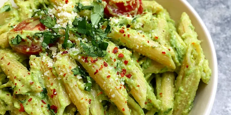 Creamy green avocado pasta in a white bowl topped with cherry tomatoes, toasted pine nuts, and fresh basil leaves